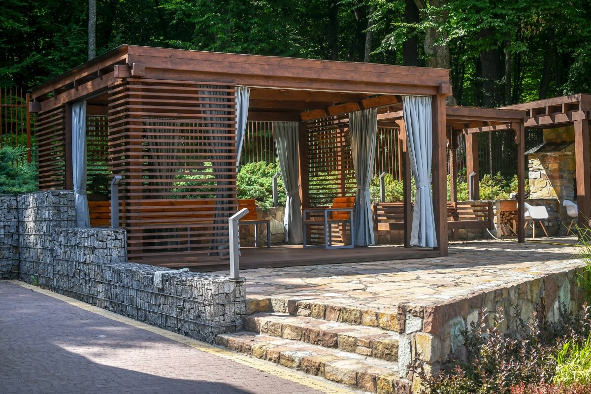 A gazebo with wooden garden blinds and gray curtains is set in a lush garden.