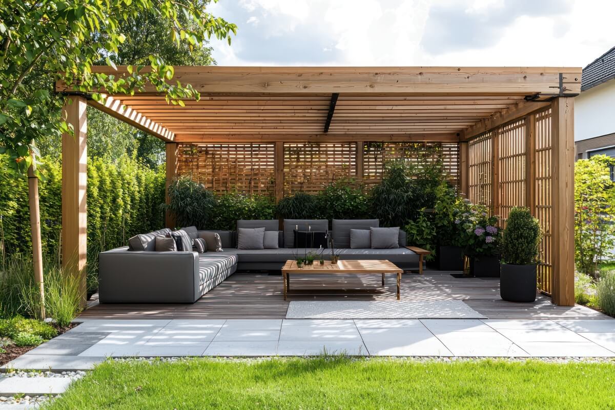 A modern patio features a comfortable seating area under a pergola with wooden terrace blinds, surrounded by lush greenery.