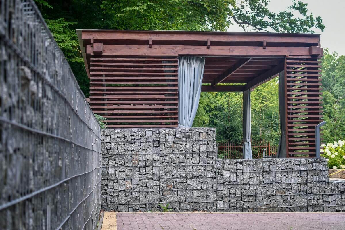 Ein Pavillon mit Vorhängen ist umgeben von Holzlamellen und einer dekorativen Steinmauer.